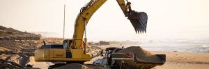 Yellow excavator raises its bucket while dump truck holds sand at the beach. Warm light background.